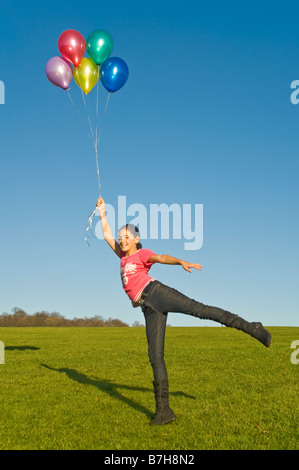 Ritratto verticale di una giovane ragazza che viene sollevato in aria da un mazzetto colorato e riempito di elio palloncini contro un cielo blu Foto Stock