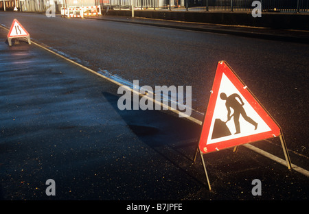 Soleggiato umido strada nero con rosso e bianco segno Roadworker e distante strada si restringe segno con opere degli ostacoli di fronte Foto Stock