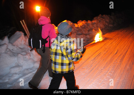 Donna adulta e giovane ragazzo uphil camminata sulla neve via illuminate da fiaccole Foto Stock