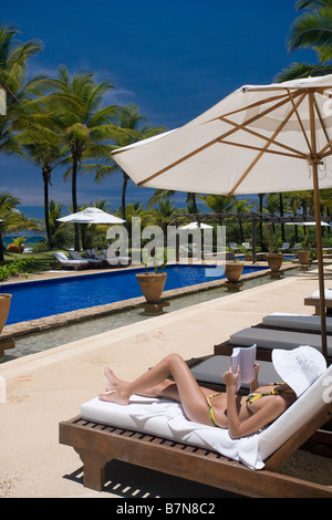 Giovane donna leggendo un libro sulla sdraio sotto ombrellone bianco accanto alla piscina nei giardini dell'hotel in Brasile Foto Stock