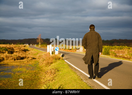 Un uomo che cammina lungo un tratto di strada nella nuova foresta, Hampshire. Regno Unito. Sole serale dietro causando lunghe ombre. Nuvole scure in anticipo. Foto Stock