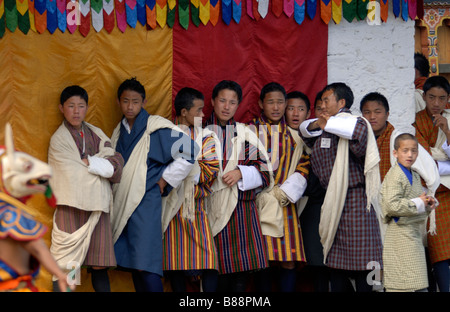 Giovani uomini bhutanesi in abito nazionale, gho, guardando danzatori mascherati nel cortile del Mongar Dzong durante il festival Mongar Foto Stock