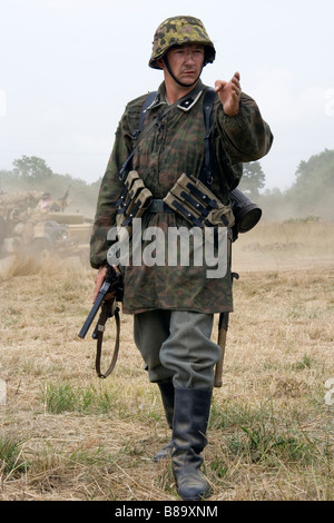 Ufficiale tedesco che indossa camouflage smock in piedi spettatore, con braccio allungato come emette ordini in un campo. Seconda rievocazione della guerra mondiale. Foto Stock