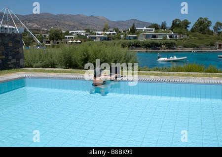 Donne che indossano cappello per il sole e il costume nero lavorando sul computer portatile in piedi in piscina, Elounda, Creta, Krētē, Grecia, Europa UE Foto Stock