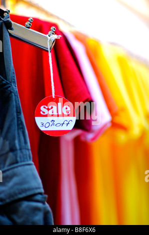 Il bianco e il rosso vendita segno appeso ad un appendiabiti di fronte a una fila di colorati T shirt Foto Stock