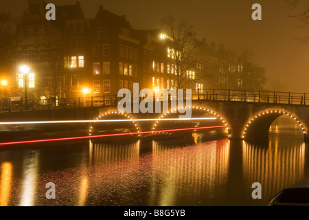 Tour in barca viaggi sotto un ponte luminoso riflesso nel canale in una nebbiosa notte a Amsterdam Paesi Bassi Foto Stock