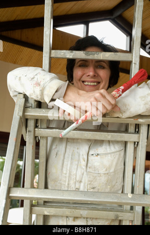 Pittore femmina in tute, strumenti in mano, sorridere mentre in piedi sulla scaletta. Foto Stock
