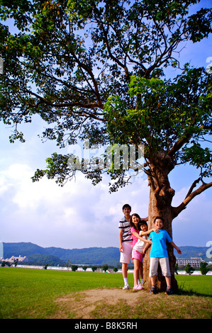 Giovane famiglia con due bambini che giocano sotto un albero alto guardando la telecamera insieme Foto Stock