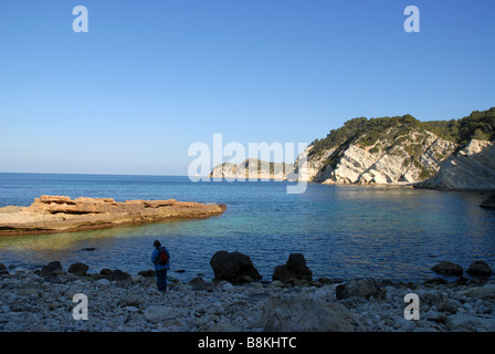 Donna che cammina sulla spiaggia rocciosa in inverno, Cap Prim dietro, Javea / Xabia, Provincia di Alicante, Comunidad Valenciana, Spagna Foto Stock