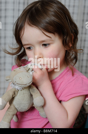 Una giovane ragazza succhia il suo pollice mentre si tiene il suo giocattolo preferito Foto Stock