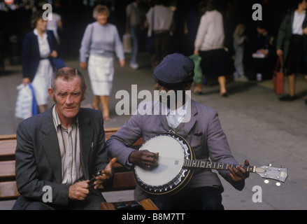 Un uomo britannico di colore che cammina con un uomo irlandese che gioca i cucchiai al centro di Manchester anni '1980 circa 1985 UK HOMER SYKES Foto Stock