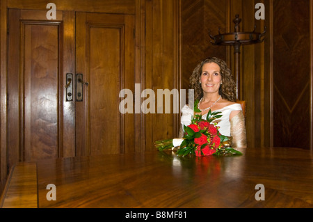 Chiudere orizzontale su ritratto di una sposa attraente con il suo bouquet nuziale di una dozzina di rose rosse seduta in una grande sala in legno Foto Stock