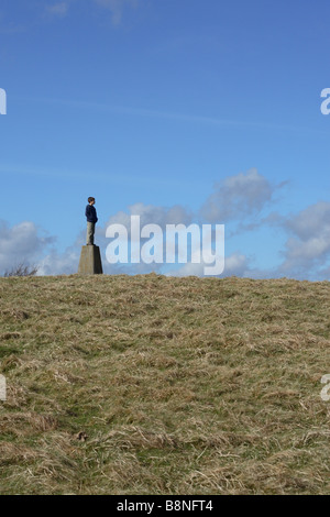 Ragazzo giovane bambino in piedi sulla cima di una collina collina punto di triangolazione marcatore in sole primaverile in Somerset Inghilterra Foto Stock