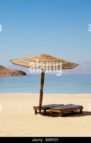 Sedie a sdraio sotto un ombrellone su una tranquilla spiaggia egiziana con la costa di Arabia Saudita visibile attraverso il Mar Rosso in distanza Foto Stock