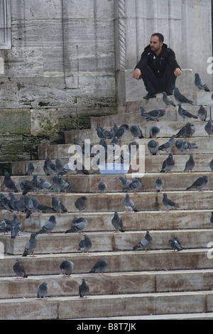 L'uomo circondato da piccioni in attesa all'ingresso della nuova moschea di Istanbul. Foto Stock