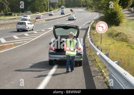 Guasto - donna che posiziona il suo segnale rosso a triangolo dietro la sua auto in avaria su una strada di svincolo su un'autostrada francese Foto Stock