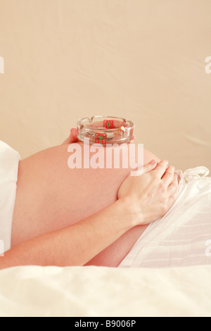 Primo piano di una piena utilizzato posacenere sul crescente bump dello stomaco di una giovane donna in stato di gravidanza nel terzo trimestre di gravidanza Foto Stock