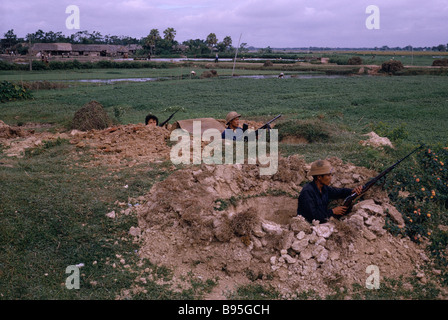 Guerra del Vietnam nord soldati vietnamiti posizionato con i fucili e mitragliatrice in scavò-outs con risaie e lavoratori dietro Foto Stock