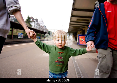 Bimbo di 2 anni a piedi nella stazione ferroviaria tenendo le mani della madre e del bambino più vecchi. Foto Stock
