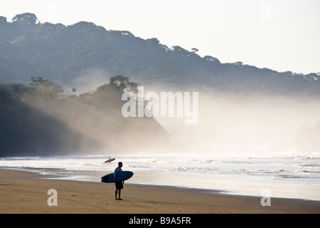 Surfers permanente sulla spiaggia con la luce del sole brilla attraverso la nebbia di mattina in Dominical, Costa Rica. Foto Stock