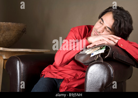 Giovane uomo contenuto in appoggio sulla sua borsa piena di denaro Foto Stock