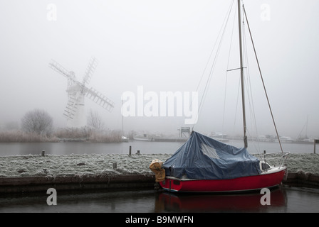 Il mulino a vento di Thurne sorge nella nebbia mentre una barca rossa chiamata richiede il primo piano Foto Stock