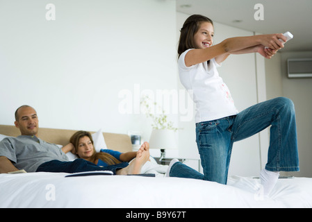 Ragazza sul letto la riproduzione del video gioco con il controller wireless, i genitori a guardare in background Foto Stock