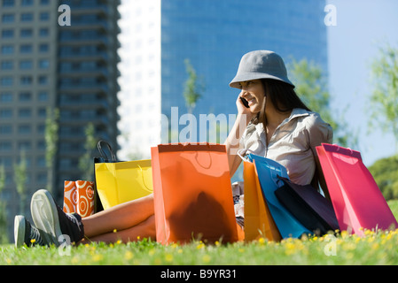 Giovane donna seduta su un terreno circondato da shopping bags, parlando al cellulare Foto Stock