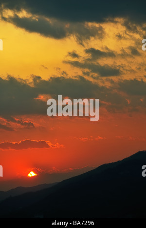 Impostazione lontano sole scompare oltre l'orizzonte avvolta in nuvole sopra le sagome delle creste di una gamma di montagna Foto Stock