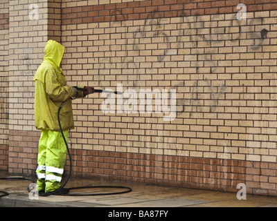 Un Lavoratore che indossa impermeabile giallo panni di pulitura a spruzzo off graffiti su un muro di mattoni con un tubo flessibile ad alta pressione Foto Stock