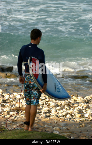 Un surfista contempla che entra in mare in possesso di una tavola da surf sotto il suo braccio in Bathsheba Beach Barbados Foto Stock