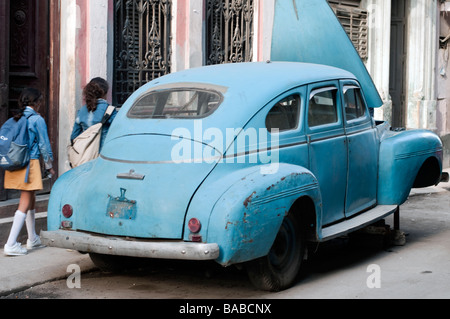 Auto antiquariato su blocchi su una strada nel centro di Havana, Cuba con due studentesse di passaggio. Foto Stock