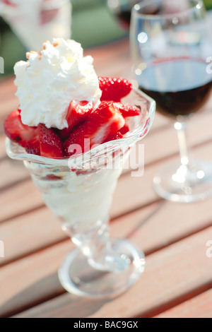 Roteato coppa di gelato guarnita con fragole e panna e a sfocare un bicchiere di vino rosso in background. Foto Stock