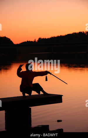 Una giovane donna in silhouette su un molo al tramonto praticare il tai chi con una spada Limousin Francia Foto Stock