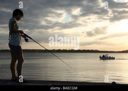 La donna la pesca su una dock sul Wilmington fiume durante il tramonto, con un motore da diporto imbarcazione in background, Savannah, Georgia Foto Stock