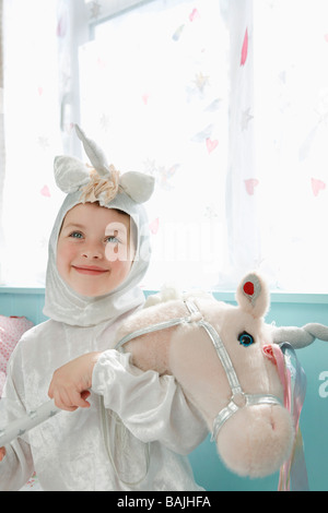 Ritratto di giovane ragazza (5-6) in costume di unicorn holding cavallo giocattolo, sorridente, interni Foto Stock