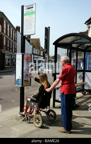 Femmina sedia a rotelle non valido utente e caregiver maschio controllare gli orari degli autobus ad una fermata del bus Foto Stock