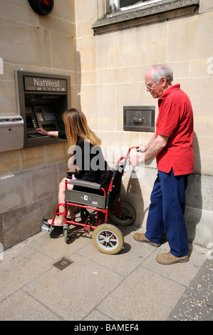 Femmina sedia a rotelle non valido utente e caregiver maschio utilizzando un banco cassa England Regno Unito Foto Stock