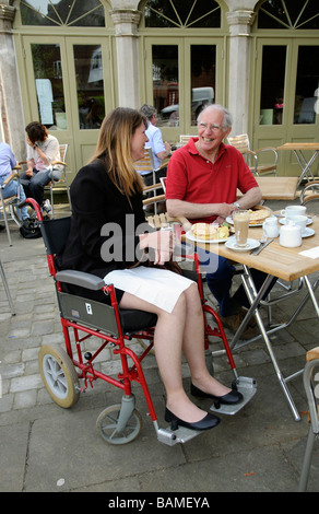 Donna seduta nella sua sedia a rotelle godendo il pranzo in un pavement cafe ristorante con il suo caregiver Foto Stock