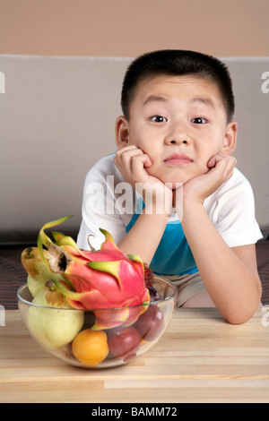 Cinese di età elementari ragazzo appoggiato su un tavolo accanto a una coppa di frutta Foto Stock