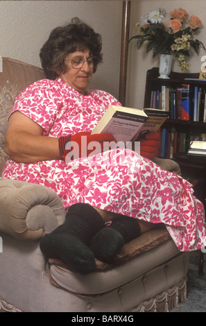 Anziani donna seduta al chiuso la lettura di un libro con i guanti e le calze su a causa di scarsa circolazione Foto Stock