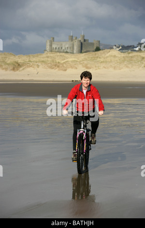 Città di Harlech, Galles. Signora escursioni in bicicletta sulla spiaggia con dune di sabbia e Harlech Castle in background. Foto Stock