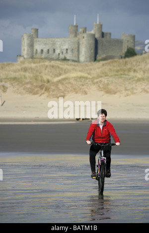 Città di Harlech, Galles. Signora escursioni in bicicletta sulla spiaggia con dune di sabbia e Harlech Castle in background. Foto Stock