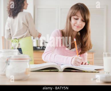 La ragazza (10-11 anni) studiare in cucina mentre la madre è la cottura Foto Stock
