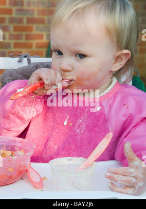 Il Toddler, 16mesi ragazza , alimentazione di se stessa e provocando un pasticcio Foto Stock