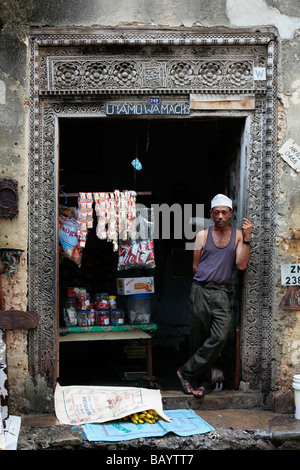 Negoziante in un portale in pietra di Zanzibar Town con gli ornati di scultura tradizionale visibile nella porta di legno Foto Stock