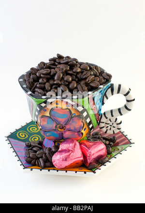 Tazza colorata e piattino traboccante di chicchi di caffè. Piattino contiene anche due Cuori di cioccolato avvolto in lamina di rosa. Foto Stock