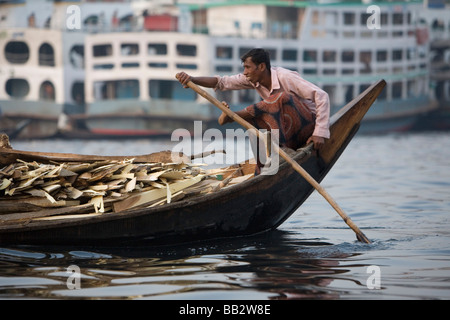 La vita quotidiana in Bangladesh; un uomo pagaie la sua barca a remi intorno al trafficato porto sul fiume Buriganga a Dhaka. Foto Stock