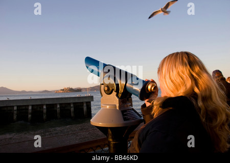 In San Francisco al Fishermans Wharf, una femmina bionda guarda attraverso un telescopio a Isola di Alcatraz con un gabbiano flying overhead Foto Stock