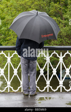 Uomo in piedi sul ponte sotto la pioggia con ombrello nero etichettato Enterprise Foto Stock